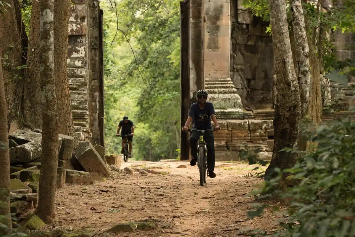 Two riders cycling through jungle ruins near Angkor.